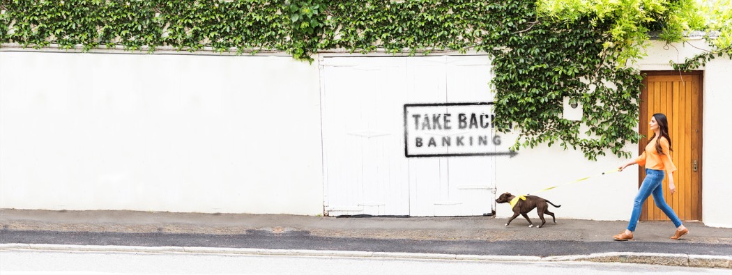 A person walking a dog along a neighborhood street with a sign reading ‘Take Back Banking,’ promoting Kasasa® checking accounts.