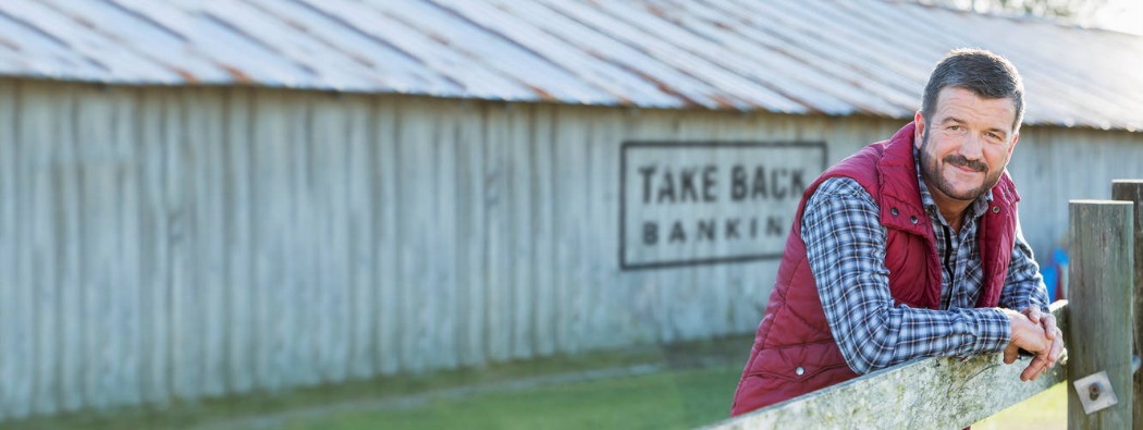 Farmer standing by a fence with a sign reading ‘Take Back Banking’ advertising Kasasa® checking accounts.
