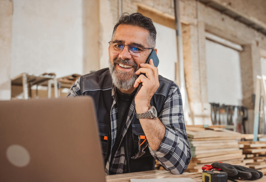 Man in a wood shop using a laptop and talking on a phone
